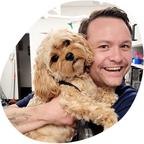 A smiling veterinarian with short brown hair hugs a small, fluffy tan dog. The man appears happy, and the dog looks at the camera with its head tilted. The background shows a home interior with shelves and various items.