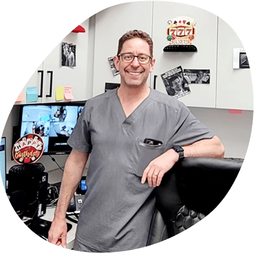 A veterinarian in gray scrubs smiles while standing in an office decorated with birthday balloons, cards, and casino-themed items. Computer monitors and cabinets with notes are visible in the background.
